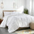 A neatly made bed with a light wood headboard features the Downlite White Goose Down Comforter | Medium Weight, white bedding, and pillows. A plant by the window, beige pouf, yellow-gray rug, nightstand lamp, and abstract art complete the scene.