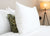 A close-up shows a neatly made bed with a Sysco Guest Supply Dream Escape Down Alternative Pillow (formerly Dream Essence) among white and green pillows. White bedding, a beige wall, and a glowing tripod floor lamp complete the tranquil scene.