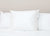 A single Keeco Best Western® Everest Pillow | Soft Polyester Pillow with a fabric tag sits upright against a white headboard and bedding, set against a plain light tan wall for a clean, cozy look.