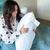 A woman with long brown hair, in a white polka dot pajama top, sits on teal bedding holding a tagged Hollander Holiday Inn® Soft and Firm Polyester Pillow Combo Pack (2 pillows), and smiles in a softly lit bedroom.