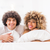 A man and woman with curly hair smile and relax in bed, holding soft Downlite Primaloft Down Alternative Pillows. Both wear white shirts, enjoying a cozy atmosphere with a bright, softly blurred background.
