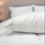 A Downlite 75/25 Waterfowl Feather/White Down Pillow by Pillow Factory rests on a neatly made bed with white bedding, while another pillow and a softly lit lamp in the background create a calm, cozy atmosphere.